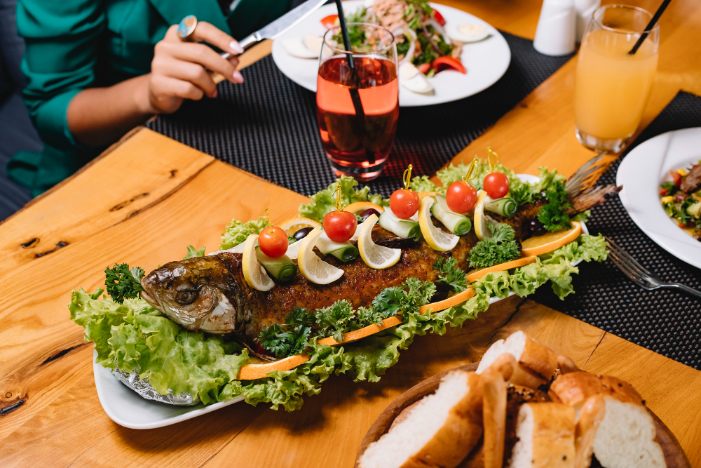 A whole grilled fish served on a platter with lemon slices, cherry tomatoes, and lettuce, accompanied by bread and a glass of juice.
