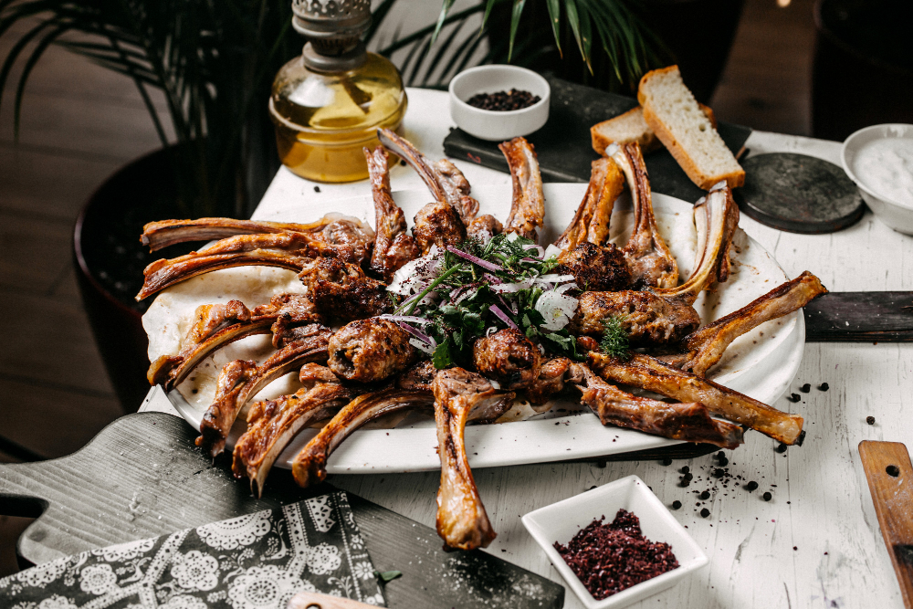 A platter of grilled lamb chops, arranged in a circular pattern and garnished with herbs and vegetables.
