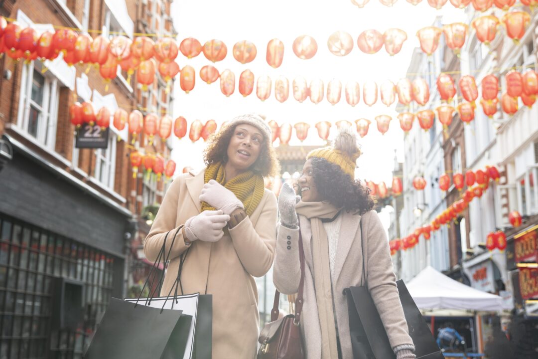 Two smiling women are walking through a lively city market in London. They are dressed casually, carrying shopping bags, and appear to be enjoying their time amid the vibrant atmosphere and market stalls.