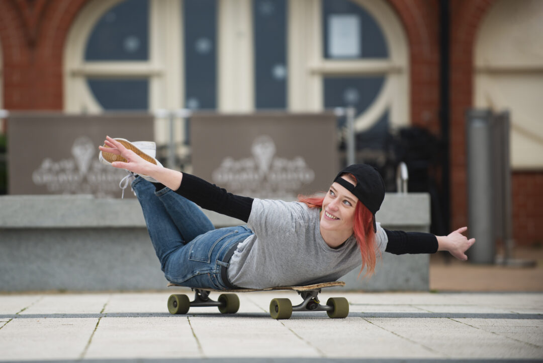 A smiling woman skateboarding outdoors in a relaxed and confident pose. She is dressed casually and the background is bright, suggesting a sunny day.