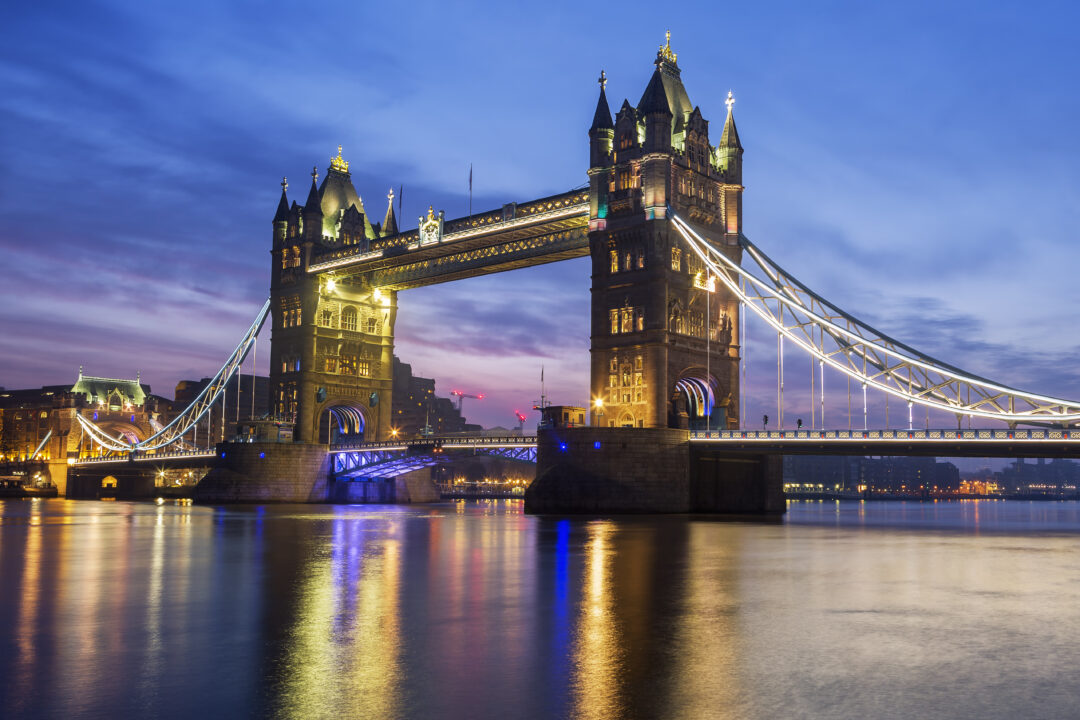 London's Tower Bridge illuminated during the evening, reflecting on the Thames River. The scene captures the bridge's iconic twin towers and suspension elements, set against a darkening sky with city lights adding warmth.